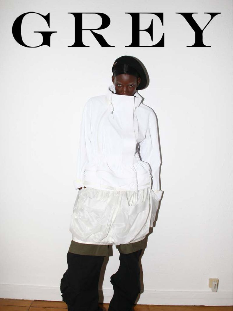 Fashion-forward portrait of a Black woman in a white jacket, shorts, and black boots, set against a stark white wall with 'GREY' typography.