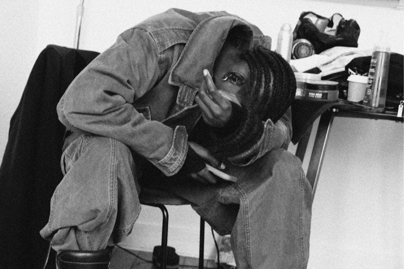 Monochrome portrait of an African American adult in a denim jacket and jeans, seated with face partially obscured by hand indoors.