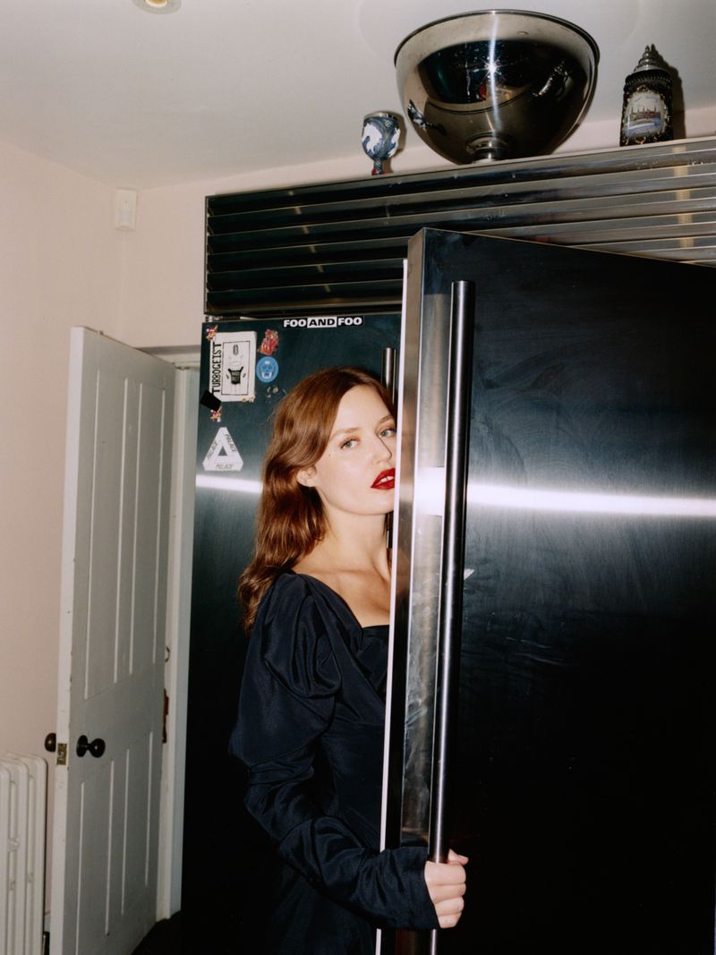 Woman in a dark dress stands holding a black refrigerator door in a brightly lit kitchen with stainless steel accents.
