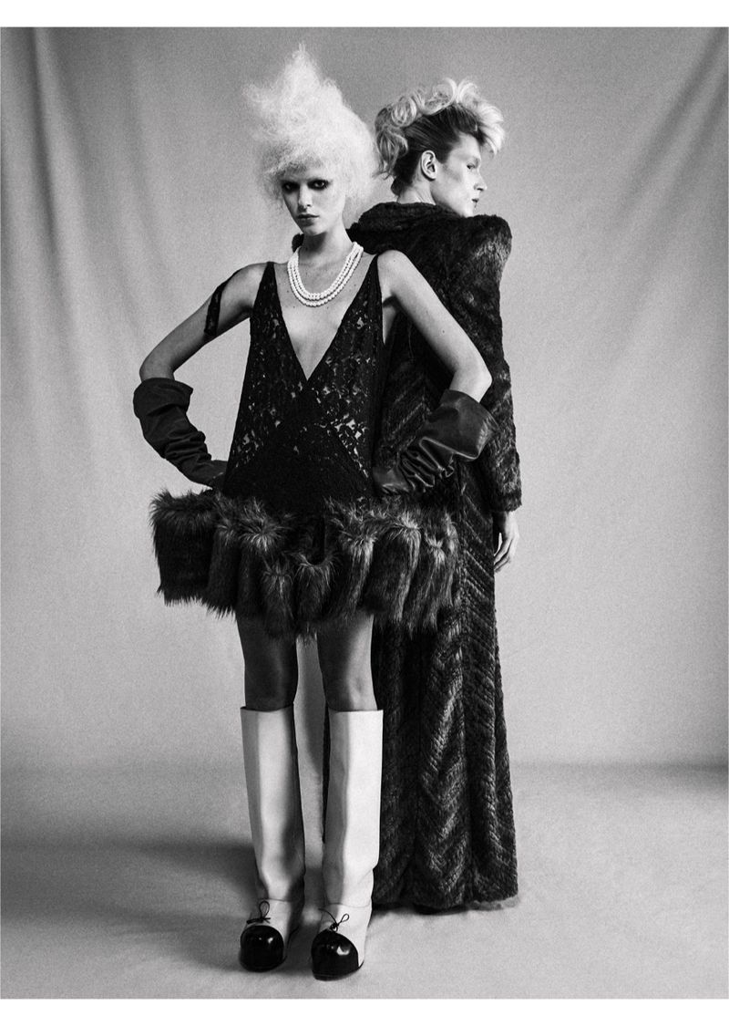 Two models pose back-to-back in a studio, showcasing avant-garde fashion with striking outfits, exaggerated hair, and bold styling.