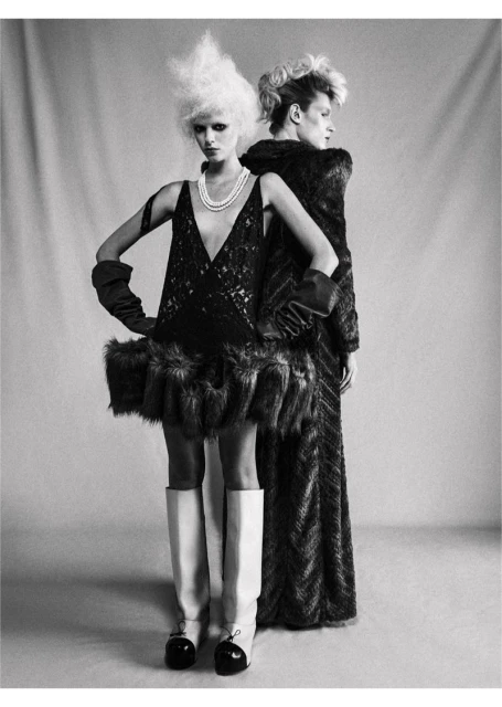 Two models pose back-to-back in a studio, showcasing avant-garde fashion with striking outfits, exaggerated hair, and bold styling.