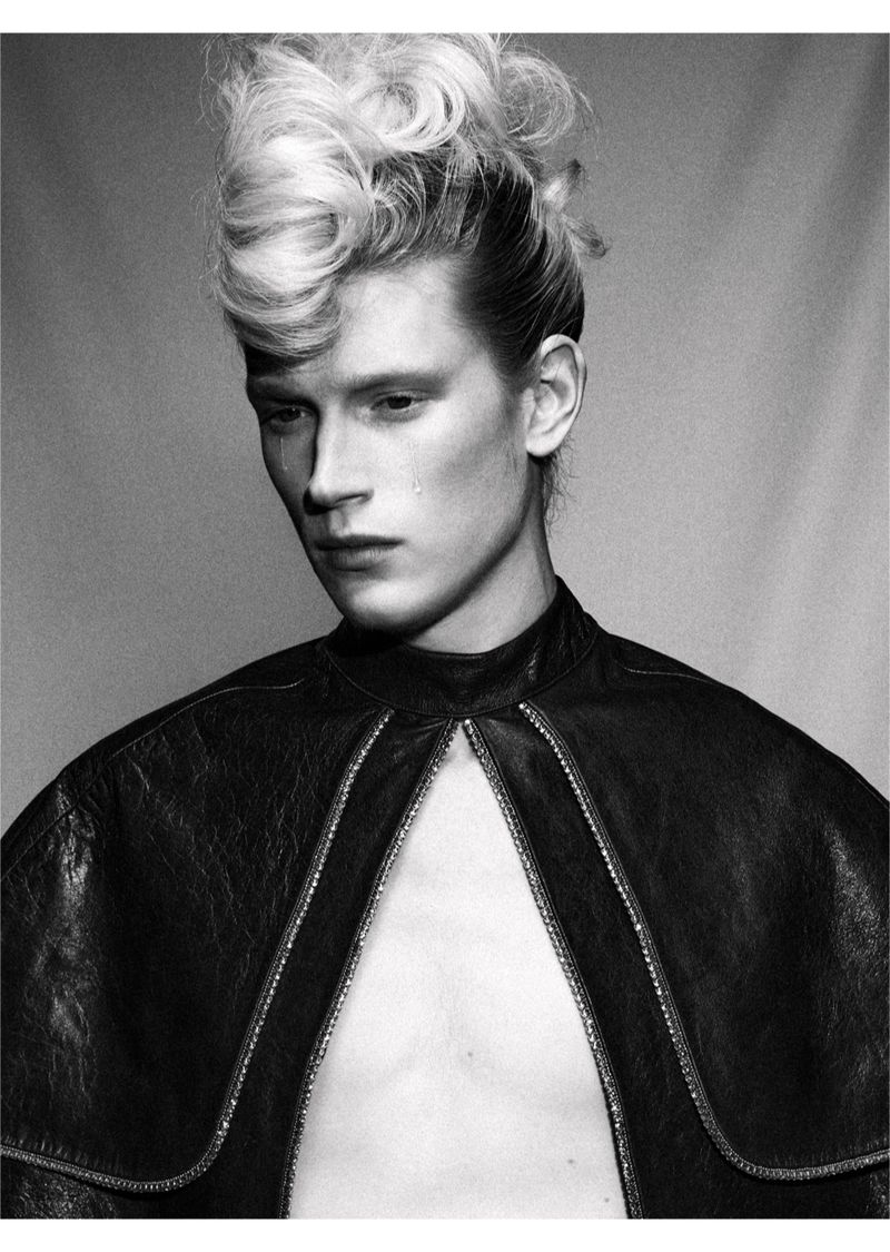 Monochrome portrait of a young adult male fashion model with a dramatic hairstyle and leather cape, gazing downward with a single tear.