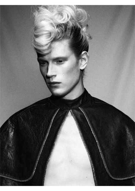 Monochrome portrait of a young adult male fashion model with a dramatic hairstyle and leather cape, gazing downward with a single tear.