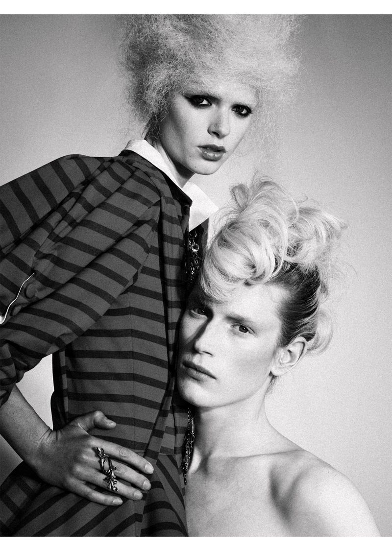 Two models pose together in a studio shot. The model in the foreground is bare-chested with stylized hair, while the other wears a striped top.