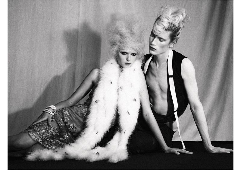 A black and white fashion shot of two adult models, a woman in a feathered shawl and dress, and a topless man in a vest, indoors.