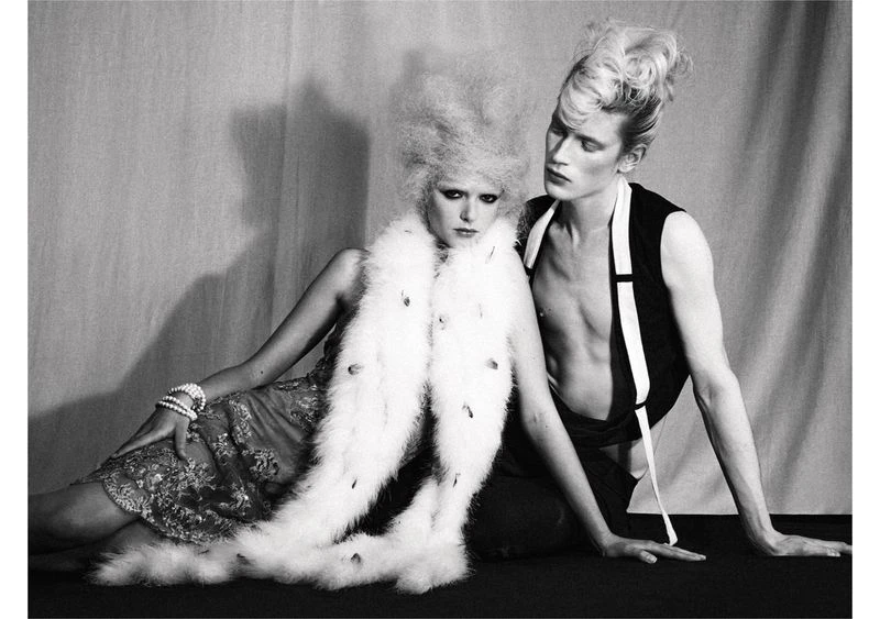 A black and white fashion shot of two adult models, a woman in a feathered shawl and dress, and a topless man in a vest, indoors.