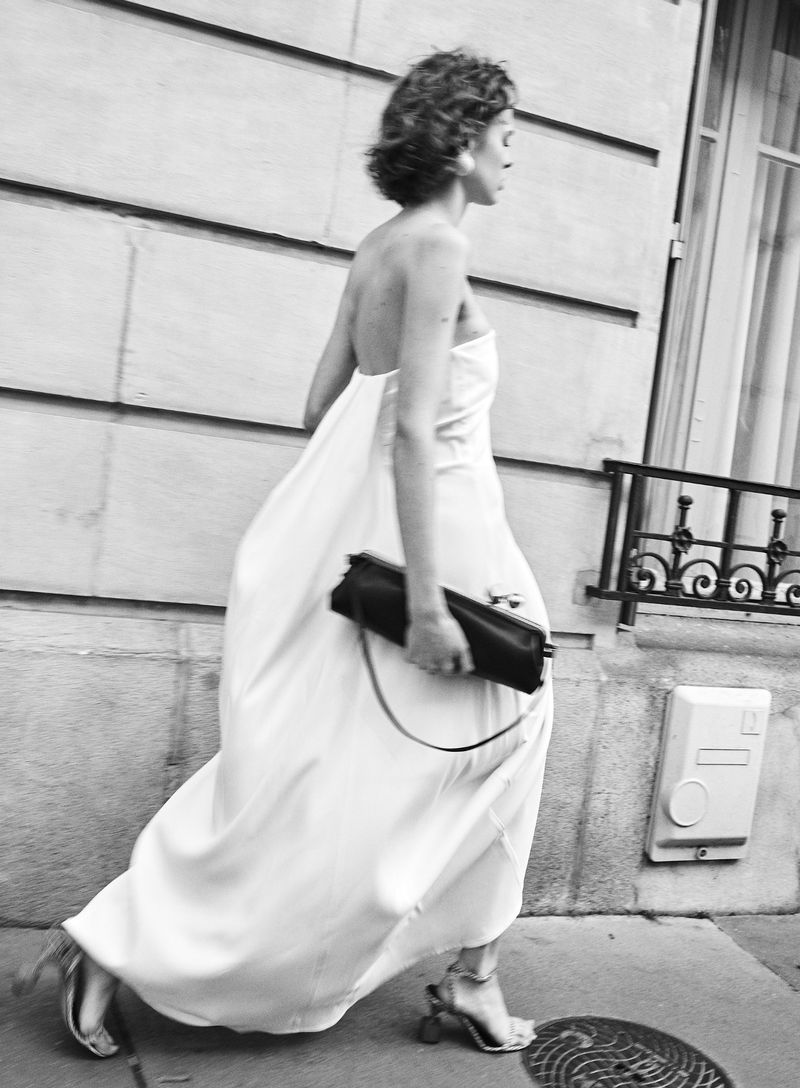 Model in a white strapless gown and black clutch walking past a stone building exterior