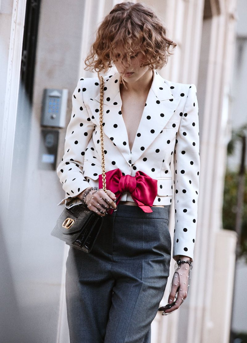 Model wearing a white polka dot jacket with a pink bow and grey trousers holding a black handbag outdoors