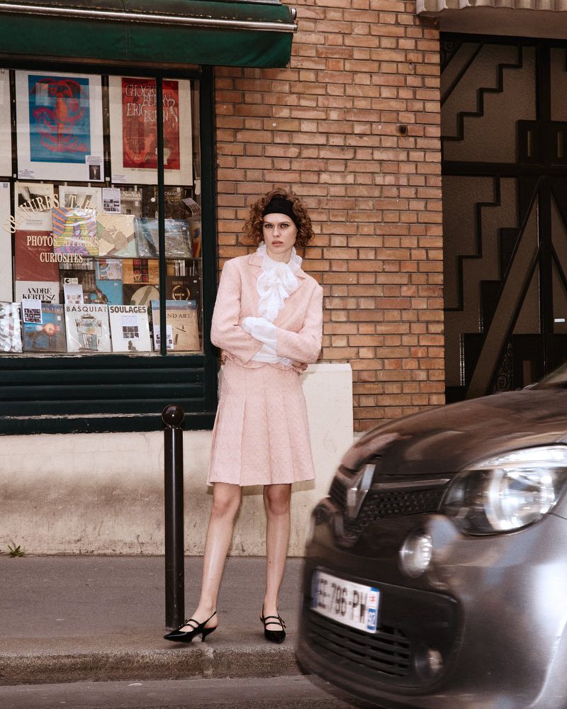 A woman in a pink suit poses on a city street near a brick building with a shop window; a gray car passes by on the right side.
