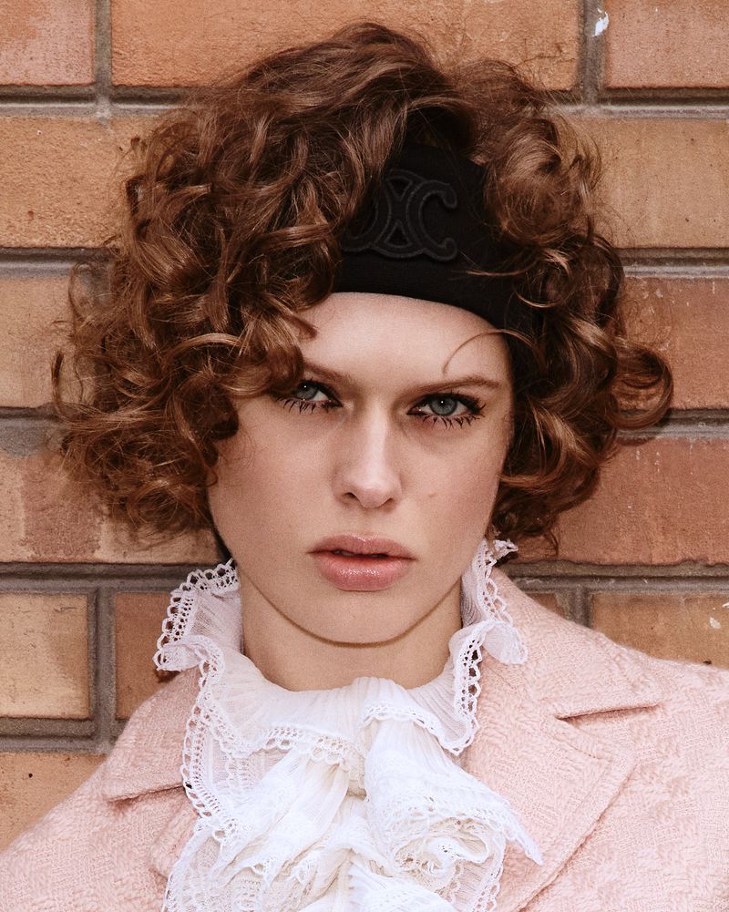Close-up portrait of a woman with curly hair and a black headband, wearing a frilled white top and a pink jacket against a brick wall.