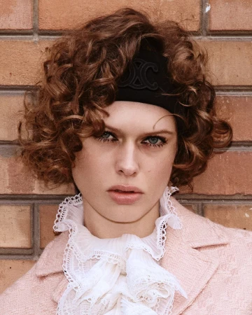 Close-up portrait of a woman with curly hair and a black headband, wearing a frilled white top and a pink jacket against a brick wall.