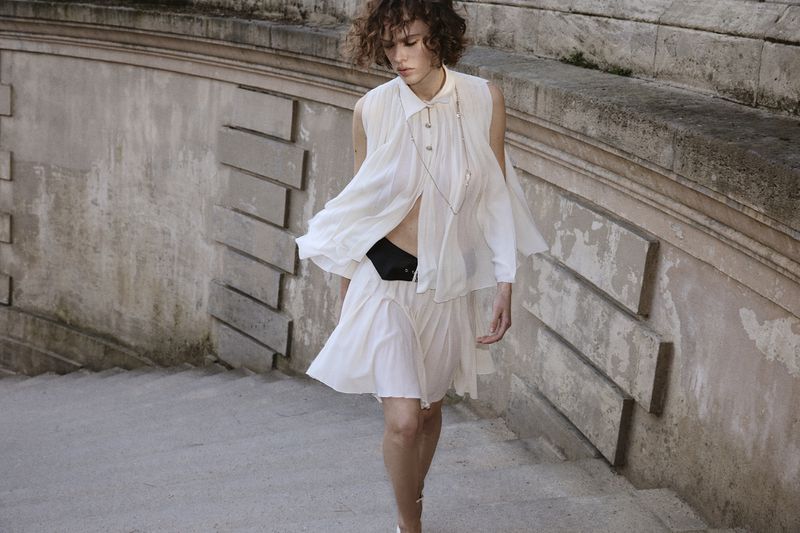 A female fashion model with curly hair poses on outdoor stairs, wearing a white layered dress and a black belt bag.