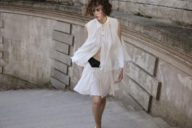 A female fashion model with curly hair poses on outdoor stairs, wearing a white layered dress and a black belt bag.