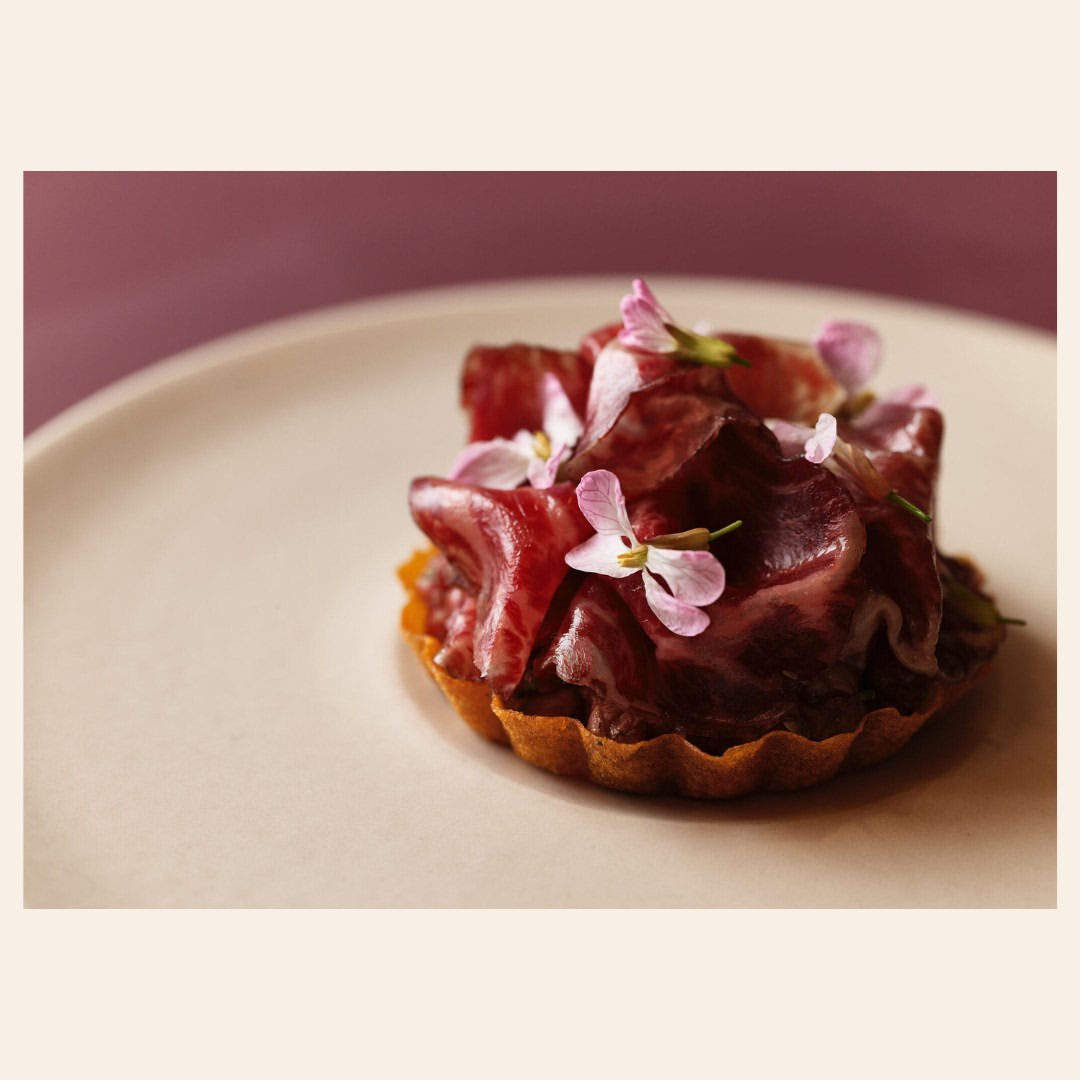 Elevated shot of an appetizer tartlet topped with deep-red thinly sliced meat and small light-purple flowers displayed on a white plate.