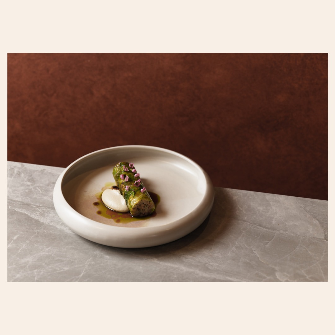 Artfully plated dish featuring a vegetable roll topped with edible flowers, served on a white plate against a brown and marble background.