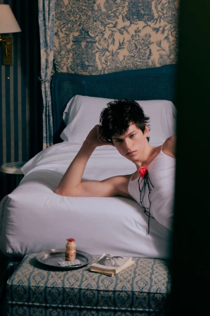 Young adult male poses in white tank top, reclining on a bed in a blue and white floral patterned room with a drink and book.