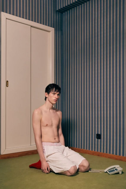 Indoor portrait of a young shirtless adult male kneeling on a cushion, featuring striped wallpaper and a vintage telephone.