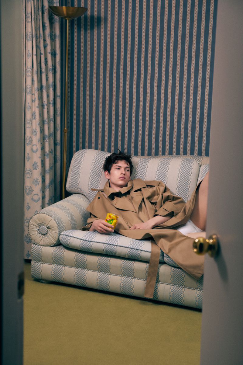 Young adult poses on a blue striped couch in a tan trench coat while holding a yellow can in an indoor studio setting.