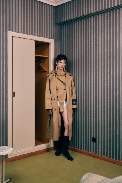 Male fashion model in a beige trench coat and tall boots poses in an indoor setting with striped wallpaper and doorway.