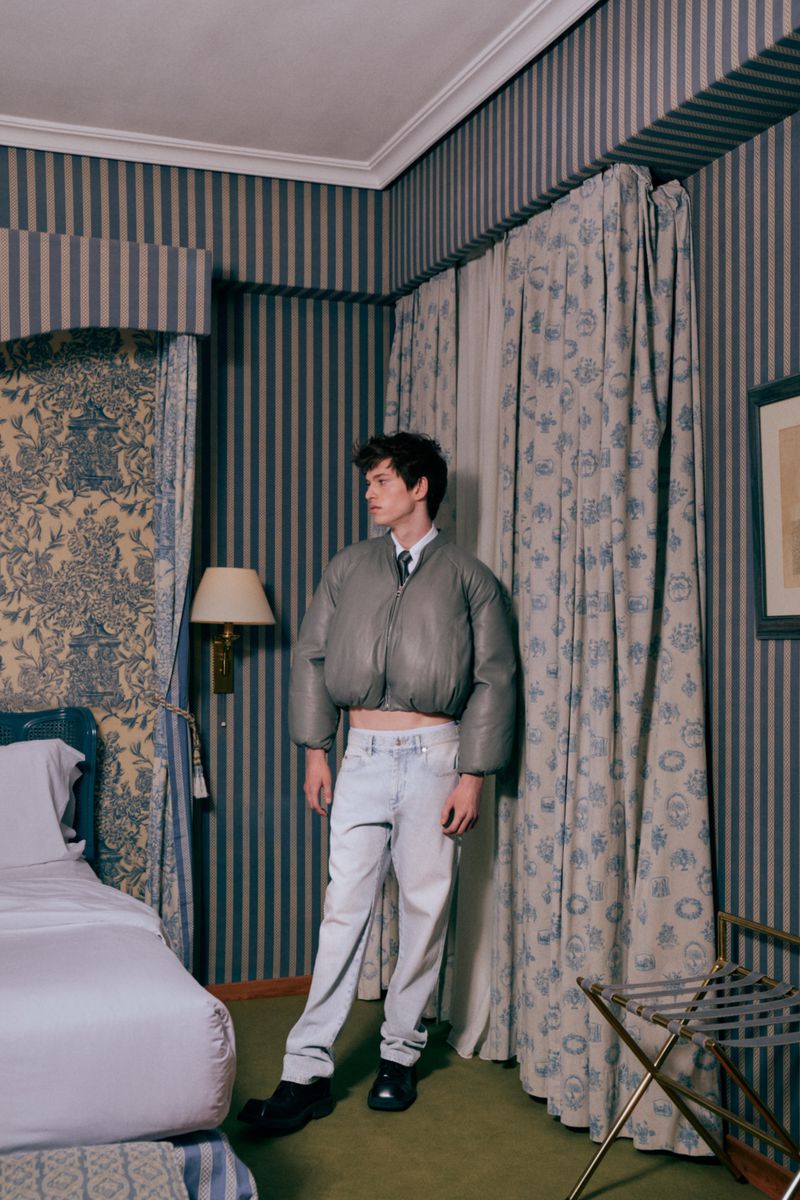 Adult male posing in a room with a bed and patterned wallpaper, wearing a cropped puffer jacket, white jeans, and black dress shoes.
