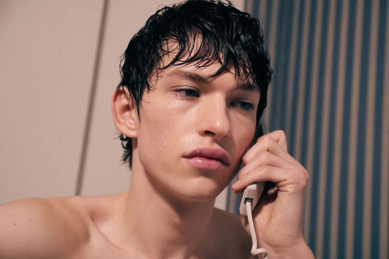 Close-up of a young adult male with wet hair holding a telephone receiver against his ear in an indoor setting with striped wall.