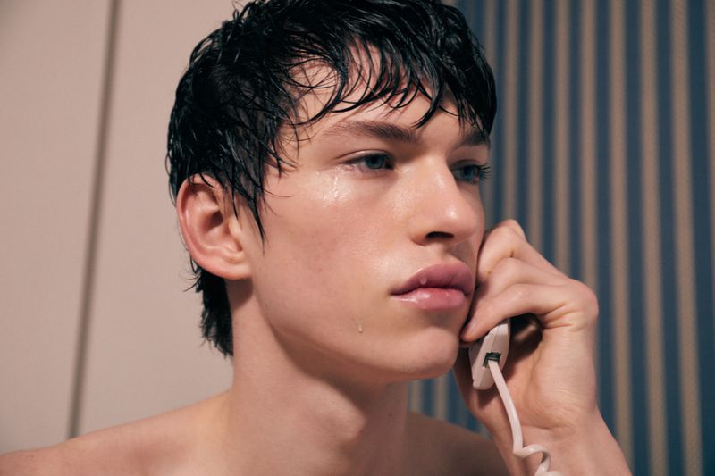 Close-up studio shot of a young adult male with wet black hair, blue eyes, and glossy lips, holding a white corded telephone to his ear.
