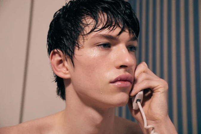 Close-up studio shot of a young adult male with wet black hair, blue eyes, and glossy lips, holding a white corded telephone to his ear.