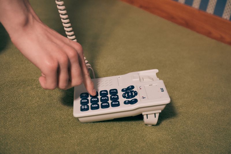 A person dials a white landline phone on a green surface, showing a close-up of the hand pressing a button with the cord visible.