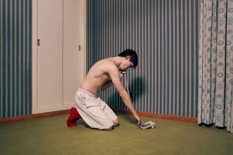 Man on his knees answers a landline phone in a room with striped wallpaper, tile floors, and a closed wardrobe. Focus is on the man.
