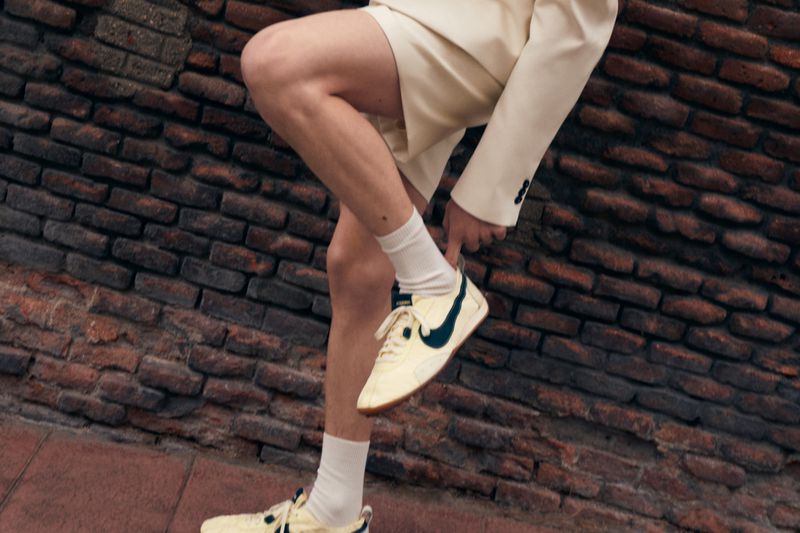 Eye-level shot of an adult wearing a cream-colored suit and cream and black trainers, standing against a textured brick wall.