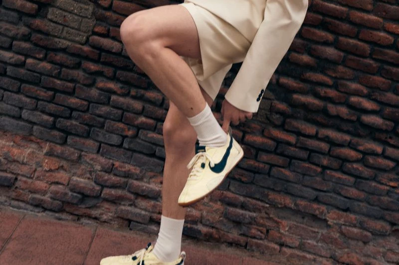 Eye-level shot of an adult wearing a cream-colored suit and cream and black trainers, standing against a textured brick wall.