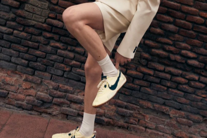 Eye-level shot of an adult wearing a cream-colored suit and cream and black trainers, standing against a textured brick wall.