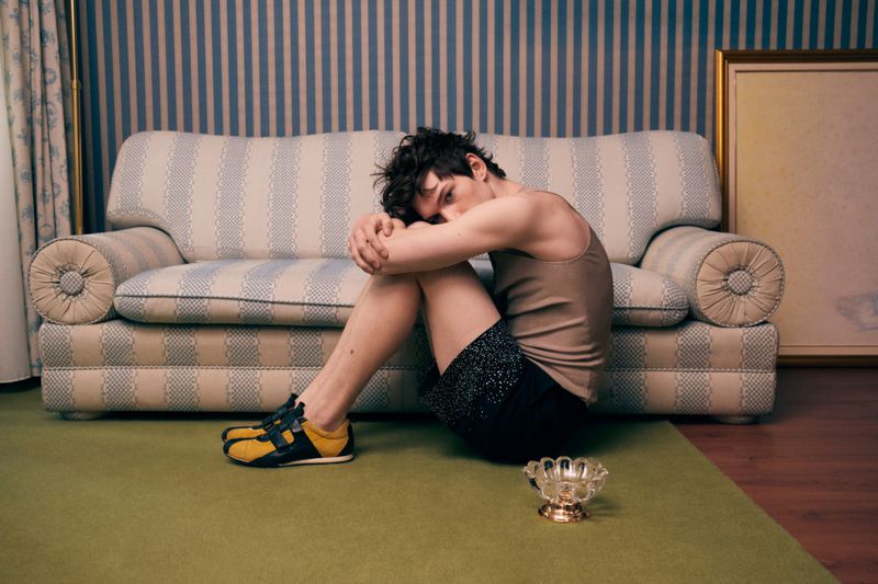 Young man in tank top sits on the floor with knees to chest, vintage trainers, a small bowl, striped wallpaper and framed art.