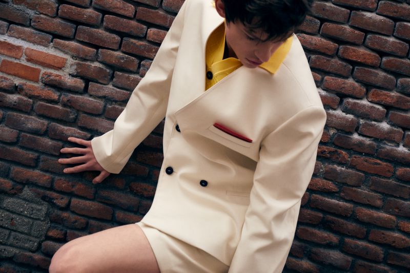 Young man in an ivory suit with yellow shirt, leaning against a textured red brick wall, in a fashion editorial shot.