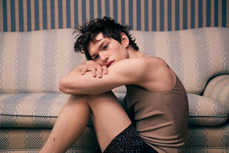 Man poses indoors sitting against a patterned sofa and striped wall, wearing a tank top and patterned shorts in soft lighting.