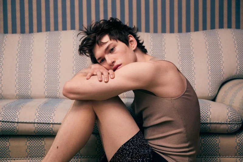 Man poses indoors sitting against a patterned sofa and striped wall, wearing a tank top and patterned shorts in soft lighting.