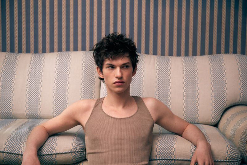 Young adult male with dark hair poses on a striped couch against a striped wallpaper background in a portrait-style shot.