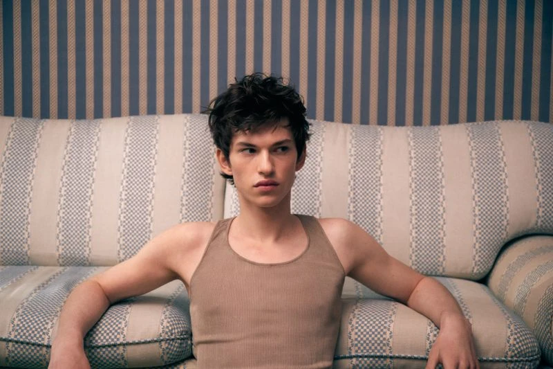 Young adult male with dark hair poses on a striped couch against a striped wallpaper background in a portrait-style shot. 
