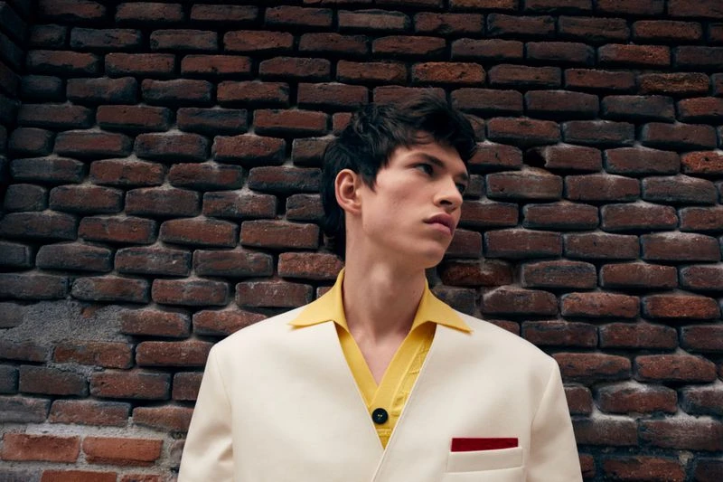 Portrait of a young adult male with dark hair wearing a yellow shirt and white jacket, posing in front of a brick wall.