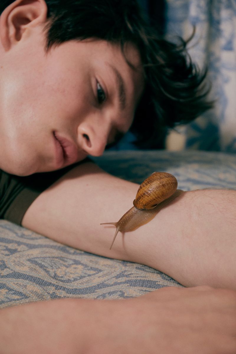 Young man lies on patterned blue bed with snail on arm, looking contemplatively at the snail in a dimly lit indoor setting.