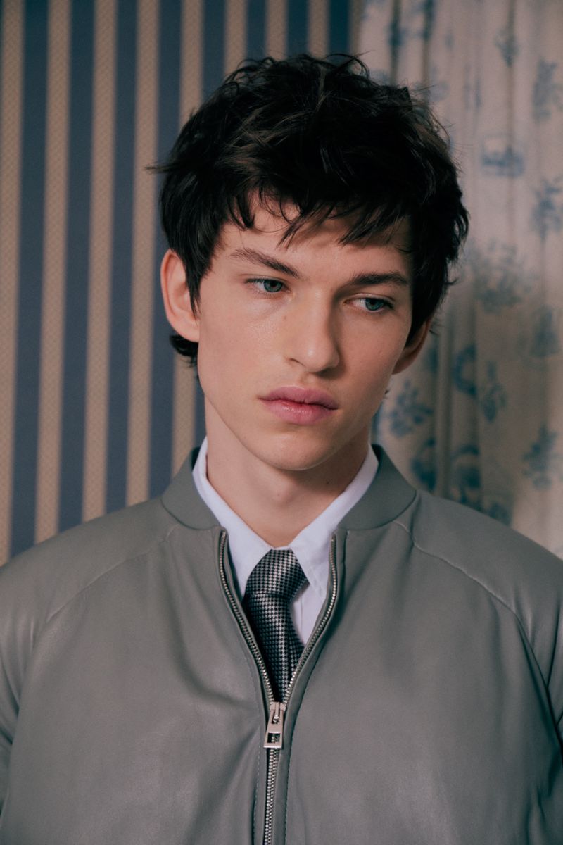 Portrait of a young male model wearing a grey bomber jacket, white shirt, and patterned tie indoors.