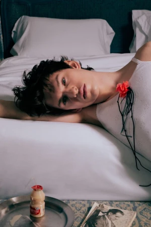 Brunette person rests on a white bed with a red flower pinned to their white tank top, bottle on a metal tray in a casual indoor scene.