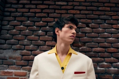 A male model wearing a cream jacket and yellow shirt against a weathered brick wall background.