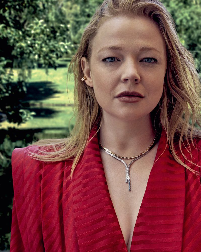 Blonde woman in red blazer poses outdoors with a green background; highlights include elegant jewelry and a fashionable, chic style.