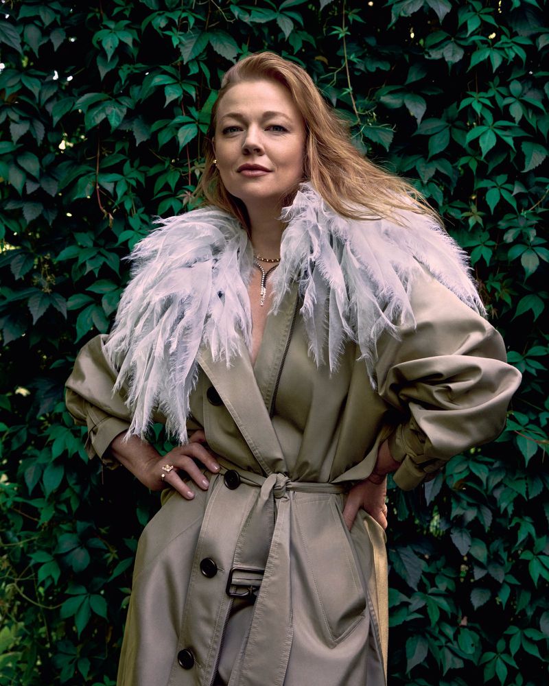 Sarah Snook wearing a belted trench coat with a feathered collar against green foliage