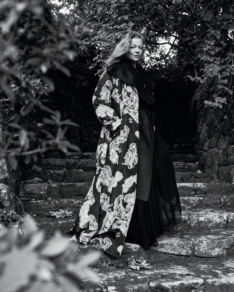 Monochrome outdoor portrait of a fair-skinned adult woman standing on stone steps wearing a patterned coat and long dark dress.