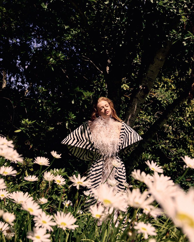A woman stands among white daisies, wearing a black and white zebra print robe with a feathered collar, in an outdoor setting.