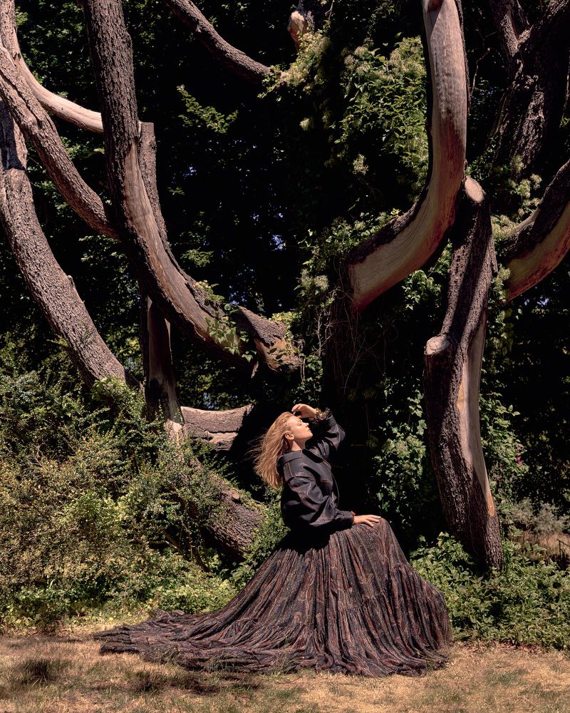 Woman in a long dress sits beneath a sprawling tree with bare branches in a lush, green landscape on a sunny day. Fashion editorial.