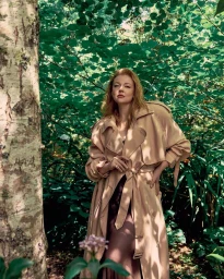 Sarah Snook in a beige trench coat standing outdoors amongst lush green foliage for a Vogue Australia editorial feature.