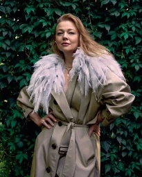 Sarah Snook wearing a belted trench coat with a feathered collar against green foliage